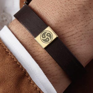 Könnte beinhalten: Ein braunes Lederarmband mit einer goldenen Metallschließe mit einem stilisierten Symbol.