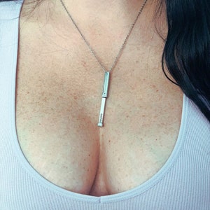 Può includere: Una collana con pendente color argento con un pendente a barra rettangolare. La parola "owned" è incisa verticalmente sul pendente. La collana è sospesa a una catena sottile.