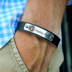 Könnte beinhalten: Schwarzes Lederarmband mit einer silbernen Platte, die mit einem Symbol und dem Text "Daddy Dom" graviert ist.