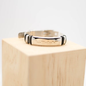 May include: A silver bracelet with a black rubber band and a silver plate engraved with braille dots.