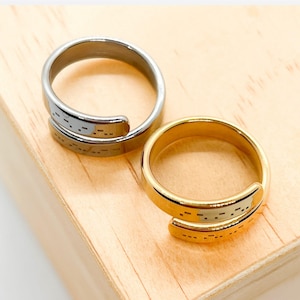 May include: Two rings, one silver and one gold, are displayed on a wooden surface. Each ring features a wrap-around design with a series of small, rectangular cutouts. The rings are simple and elegant, suitable for everyday wear.