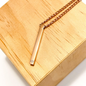 May include: Rose gold bar necklace with a delicate chain. The pendant has a small infinity symbol engraved on it. The necklace is displayed on a light brown wooden surface.