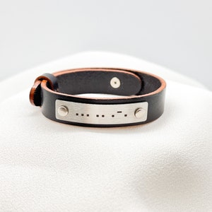 May include: Black leather bracelet with a silver metal plate. The plate has Morse code symbols. The bracelet has a brown interior and a buckle closure. The bracelet is approximately 0.75 inches wide.