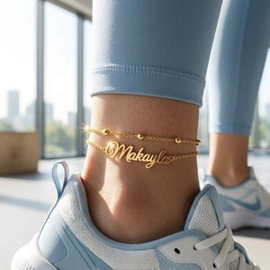 Peut inclure: Une chevillère dorée à double chaîne ornée de petites perles et d'une plaque centrale portant le prénom Makayla en écriture cursive, portée autour de la cheville au-dessus d'une basket de sport blanche et bleu clair.
