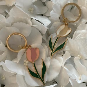 May include: Two tulip-shaped keyrings with gold-coloured rings. One keyring features a pink and light pink tulip with green leaves, and the other has a white and cream tulip with green leaves. The keyrings are set against a backdrop of white flower petals.
