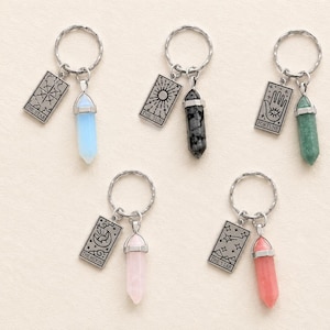 May include: Five silver-toned metal keychains, each featuring a split ring, a rectangular charm with engraved tarot-style imagery and text, and a pointed, faceted crystal pendant in light blue, black, green, pale pink, or coral colours.
