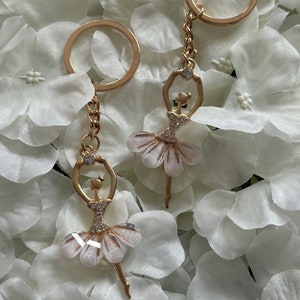 May include: Two gold-tone ballerina keyrings with white sparkly tutus. The ballerinas are holding their arms up in a graceful pose.