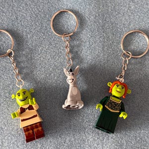 May include: Three metal keychains featuring plastic character figures. One keychain features a green ogre in a tan tunic, one features a grey donkey, and one features a green ogre with orange hair wearing a dark green dress with gold floral patterns.