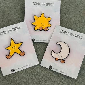 May include: Three enamel pin badges on individual backing cards labelled ENAMEL PIN BADGE and BOBBLEANDBOOGIFTS. The pins feature a yellow sun, a yellow star, and a white crescent moon, each with a smiling face and small brown boots.
