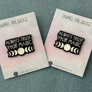 May include: Two black enamel pins on white backing cards. Each pin features the text "ALWAYS TRUST YOUR MAGIC" in white lettering above a series of white moon phases. The backing cards are labelled "ENAMEL PIN BADGE" and "BOBBLEANDBOOGIFTS".