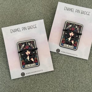 May include: Two rectangular enamel pins on white backing cards labelled ENAMEL PIN BADGE. Each pin features a tarot card illustration of a person in a wide-brimmed hat with flowers, holding a black cat, with the text THE WITCH at the bottom.