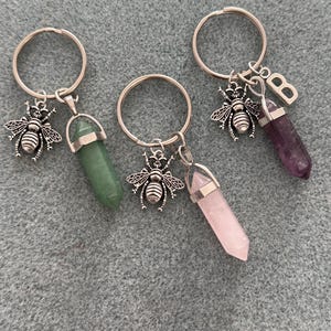 May include: Three keyrings with silver rings and bee charms. Each keyring has a different coloured crystal pendant: green, pink, and purple. The purple crystal has a silver letter "B" charm.