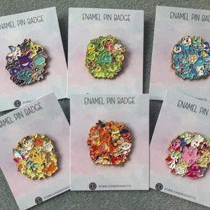May include: Six circular enamel pin badges attached to individual white display cards labelled ENAMEL PIN BADGE. Each pin features a cluster of colourful cartoon creatures in various shades of purple, green, blue, yellow, orange, and pink, with gold-toned metal outlines.