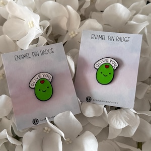 May include: Two enamel pin badges featuring a smiling green olive with the text "OLIVE YOU" above it. The pins are on cards that read "ENAMEL PIN BADGE". The background is white flowers.