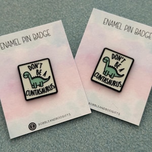 May include: Two rectangular enamel pins featuring a light green cartoon dinosaur on a cream background. The text reads DON'T BE A CUNTASAURUS in black lettering. Each pin is attached to a white backing card labelled ENAMEL PIN BADGE and BOBBLEANDBOOGIFTS.