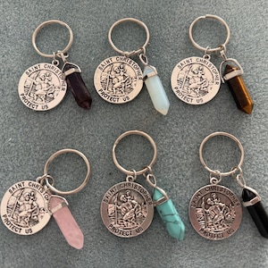 May include: Six silver keyrings with a silver charm featuring the image of Saint Christopher and the text "SAINT CHRISTOPHER PROTECT US". Each keyring has a different coloured crystal attached to it: amethyst, opal, tiger's eye, rose quartz, turquoise, and black onyx.
