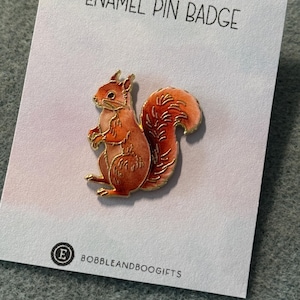 May include: An enamel pin badge shaped like a squirrel with orange and red colouring and gold metal outlines. The pin is attached to a white display card featuring the text ENAMEL PIN BADGE at the top and BOBBLEANDBOOGIFTS at the bottom.