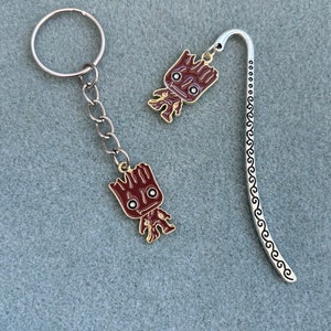 May include: A silver bookmark with a decorative design and a small, brown, cartoon character charm. The charm is attached to a silver key chain.