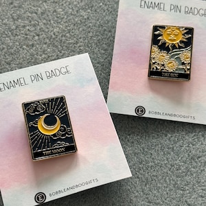 May include: Two enamel pin badges on cards. One shows a yellow crescent moon and stars on a black background with the text "THE MOON". The other has a yellow sun and flowers with the text "THE SUN". Both have gold borders.
