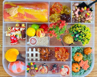 May include: A clear plastic organizer box filled with autumn-themed sensory play items, including a yellow and orange dough log, miniature scarecrows, plastic pumpkins, colorful leaves, small trees, a coffee cup charm, and turkey figurines.