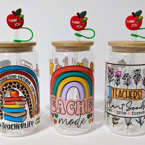 May include: Three clear glass tumblers with lids and straws. Each tumbler has a different design. One has a rainbow with the text "Teacher Mode", one has a rainbow with the text "#TeacherLife", and one has flowers with the text "Teachers Plant Seeds that grow forever".