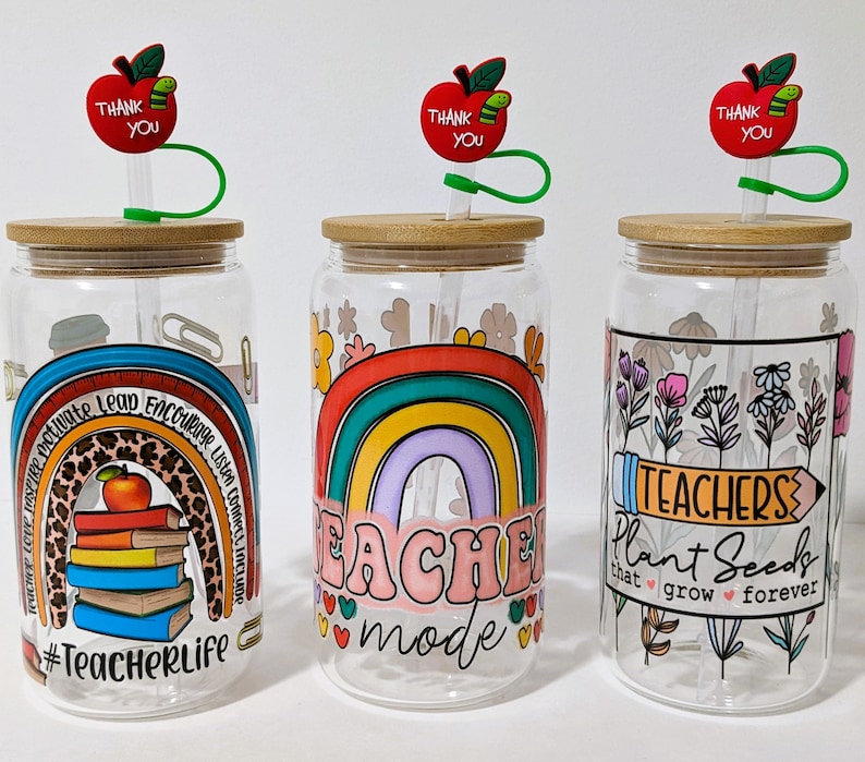 May include: Three clear glass tumblers with lids and straws. Each tumbler has a different design. One has a rainbow with the text "Teacher Mode", one has a rainbow with the text "#TeacherLife", and one has flowers with the text "Teachers Plant Seeds that grow forever".
