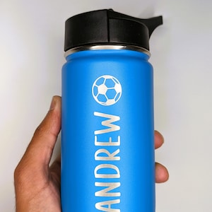 May include: A blue stainless steel water bottle with a black lid and a carrying handle. The bottle features a soccer ball design and the name "ANDREW" engraved on the side.
