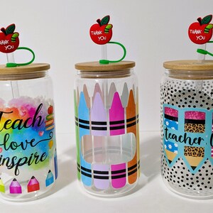 May include: Three clear glass jars with bamboo lids and green straws. Each jar has a different design: one with a rainbow watercolor background and the text "Teach love inspire", one with colorful crayons, and one with black and white polka dots and the text "teacher life". Each jar has a red apple with a green leaf and the text "Thank You" on top.