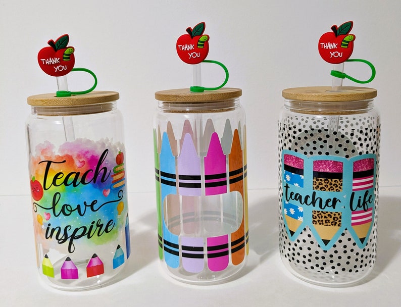 May include: Three clear glass jars with bamboo lids and green straws. Each jar has a different design: one with a rainbow watercolor background and the text "Teach love inspire", one with colorful crayons, and one with black and white polka dots and the text "teacher life". Each jar has a red apple with a green leaf and the text "Thank You" on top.