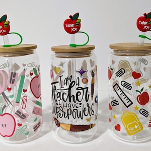 May include: Three clear glass tumblers with bamboo lids and red apple straws. Each tumbler has a different design featuring school supplies, hearts, stars, and the text "I am a teacher I have superpowers".
