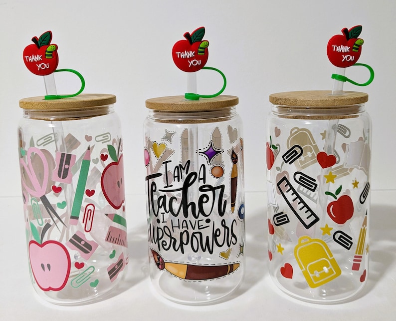 May include: Three clear glass tumblers with bamboo lids and red apple straws. Each tumbler has a different design featuring school supplies, hearts, stars, and the text "I am a teacher I have superpowers".