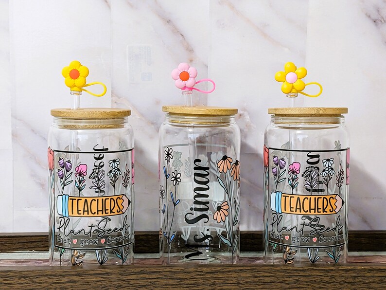 May include: Three glass jars with bamboo lids and colorful flower straws. Each jar has a different design with floral patterns and text. The text on the jars reads "Teachers Plant Seeds That Grow Forever", "Ms. Simar", and "Teachers Plant Seeds That Grow Forever".
