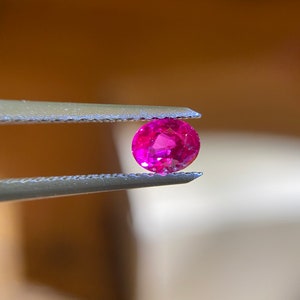 May include: A vibrant pink gemstone held in tweezers. The gemstone is round and has a smooth, polished surface.
