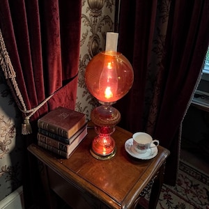 May include: An antique orange glass lamp with a round shade and ornate base sits on a wooden nightstand. Beside the lamp are a stack of old books and a teacup with a floral design. Red curtains frame the scene.