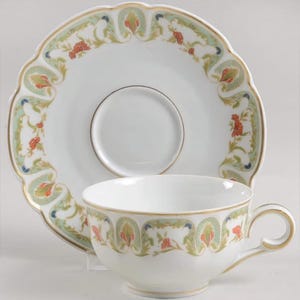 May include: A white porcelain teacup and matching saucer featuring a scalloped edge and a decorative border pattern of green leafy scrolls, small orange floral accents, and tiny blue dots, all finished with a thin gold trim along the rims.