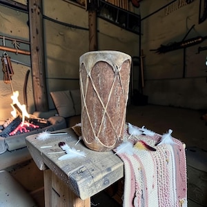 May include: A cylindrical drum with a natural wood grain body and a light-colored rawhide head secured by a crisscross lacing pattern made of thin cord. The drum is positioned upright, featuring a textured bark-like exterior surface.