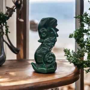May include: A glossy, dark green ceramic sculpture of a stylized figure with a spiral design. The sculpture is placed on a wooden table, with a blurred background of a window and greenery. The figure has a unique, abstract design.