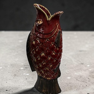 May include: A ceramic vase in the shape of a fish, with a deep red glaze and textured scales. The fish has a wide open mouth and a dark brown base and fins. The vase is approximately 10 inches tall.