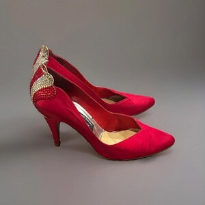 May include: A pair of red satin pointed-toe pumps featuring a mid-height stiletto heel. The back of each shoe is adorned with a decorative wing-shaped embellishment covered in sparkling clear and red crystals, with a branded interior insole.
