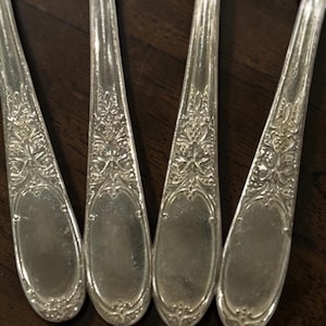 May include: Close-up of four silver spoons with ornate floral and scrollwork detailing. The spoons are arranged closely together, showcasing the intricate patterns on the handles and bowls. The spoons are made of a shiny, reflective metal.