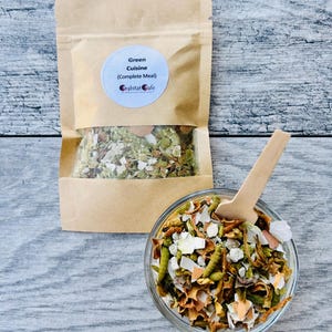May include: A clear glass bowl and a small wooden spoon filled with a complete meal. A brown paper bag with a clear window contains the same meal. The bag has a label that reads "Green Cuisine (Complete Meal) habitat cafe."