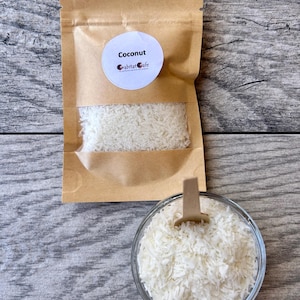 May include: A kraft paper bag and a clear glass bowl filled with shredded coconut. The bag has a circular label that reads "Coconut" and "Crabitat Cafe". A small wooden scoop rests in the bowl of coconut.
