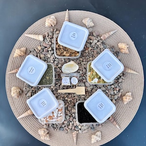 May include: A circular arrangement of small, clear containers with white lids, each labeled with text. The containers are filled with various materials, including dried leaves and black castings. The arrangement is on a woven mat surrounded by seashells and small stones.