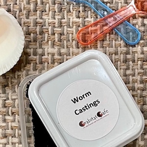 Op de afbeelding: Een close-up van een witte plastic container met het label "Worm Castings" met een zwarte substantie erin. Twee doorzichtige plastic lepels, &eacute;&eacute;n blauw en &eacute;&eacute;n oranje, kruisen elkaar boven de container. De achtergrond is een geweven stof.