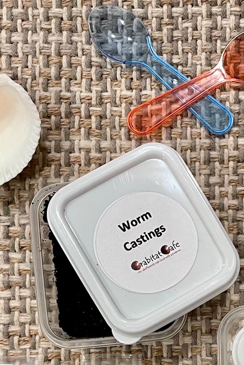Op de afbeelding: Een close-up van een witte plastic container met het label "Worm Castings" met een zwarte substantie erin. Twee doorzichtige plastic lepels, &eacute;&eacute;n blauw en &eacute;&eacute;n oranje, kruisen elkaar boven de container. De achtergrond is een geweven stof.