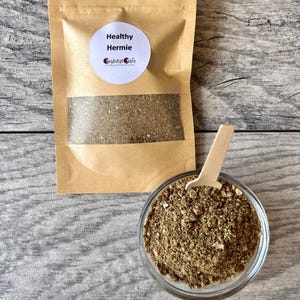 May include: A brown paper bag with a clear window, labeled "Healthy Hermie" with a logo. A small glass bowl and wooden spoon contain a brown granular substance. The background is a light gray wood grain.