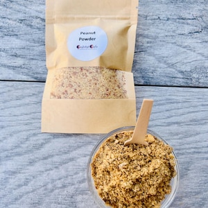 May include: A tan paper pouch labeled Peanut Powder by Grabitat Cafe containing a coarse beige and brown powder, accompanied by a clear glass bowl filled with the same textured peanut powder and a small wooden spoon inserted into the center.