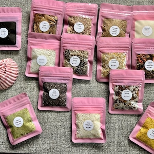 May include: A collection of small pink pouches filled with various ingredients, including seeds, powders, and dried items. Each pouch has a white label with text. A seashell is also present.