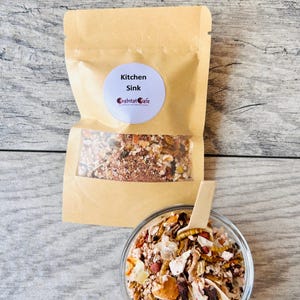 May include: A clear glass bowl and a kraft paper bag filled with a mixture of dried food items. The bag has a clear window and a label that reads "Kitchen Sink" and "Crabitat Cafe". A small wooden spoon rests in the bowl.
