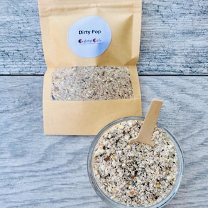 May include: A clear glass bowl and a small wooden scoop filled with a granular mixture. A brown paper bag with the text "Dirty Pop" and "habitat cafe" is behind the bowl. The mixture appears to be a blend of various grains and spices.